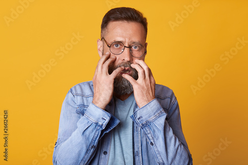 Scared man in panic, dressed in denim jacket, gray shirt and spectacles, feels fear, bits her nails, male watches horror film, model posing isolated over yellow studio background. Human emotions. © sementsova321