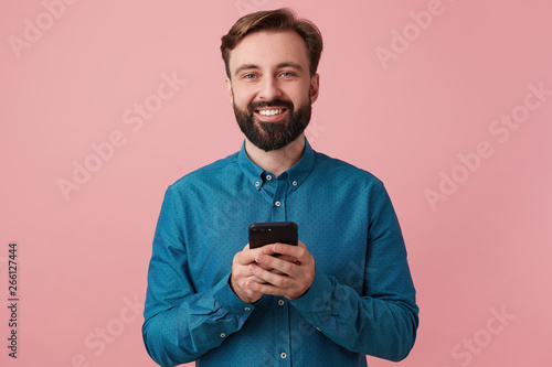 Foto Portrait of a contented attractive bearded young man, wearing a denim shirt, holding a smartphone in his hands, smiling widely and looking at the camera isolated over pink background