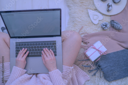 Woman's hands typing on laptop keyboard. Study and work online. Warm pink sweater, winter, home comfort and relax. Workspace with laptop, girl's hands, cake, cristmas gifts on white blanket.