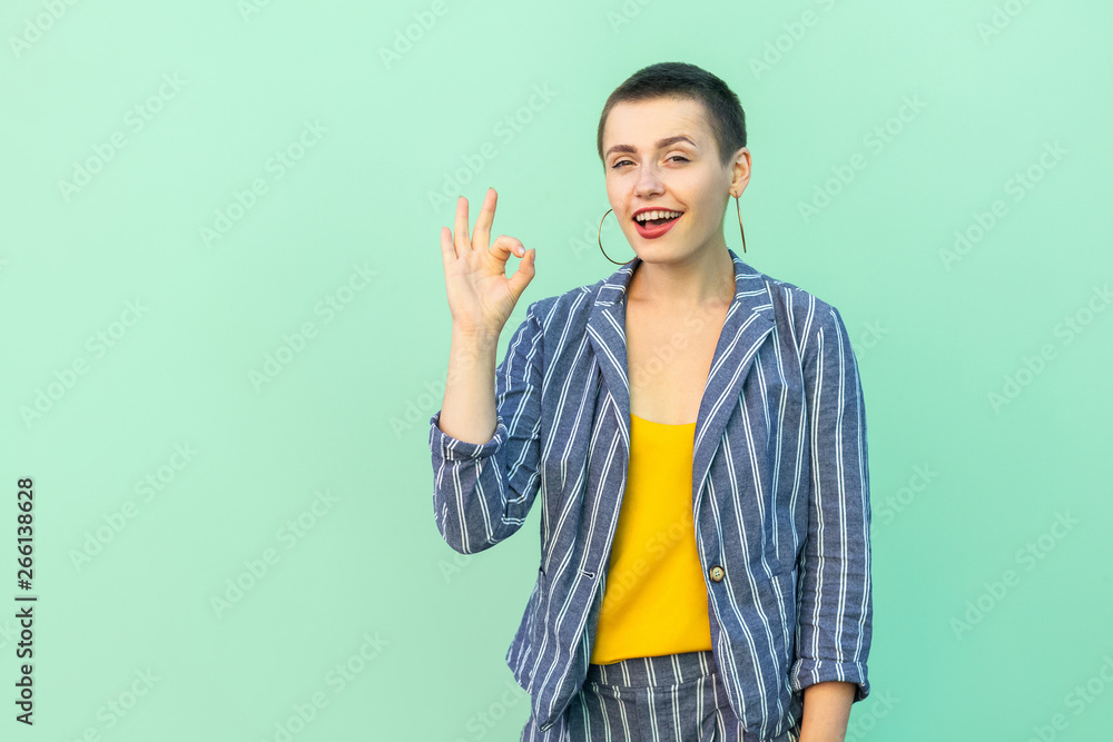 Portrait of funny handsome beautiful short hair young stylish woman in casual striped suit standing with Ok sign and looking at camera with toothy smile. studio shot isolated on light green background Portrait of funny handsome beautiful short hair young stylish woman in casual striped suit standing with Ok sign and looking at camera with toothy smile. studio shot isolated on light green background