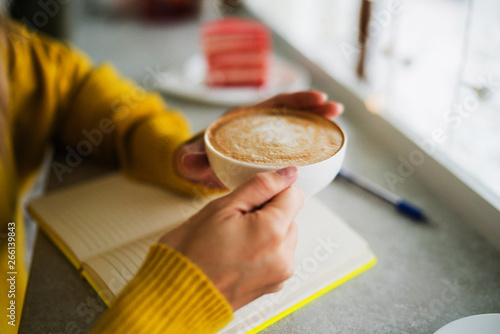 Cup of cappuccino in woman hands with cake in cafe. Hot drink. Cozy coffee shop atmosphere in autumn or winter. Autumn or winter relax concept. Warm vintage toning. Selective focus.