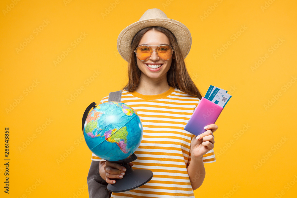 Cheerful girl holding globe in one hand and passport with flight tickets in other, isolated on yellow background Cheerful girl holding globe in one hand and passport with flight tickets in other, isolated on yellow background