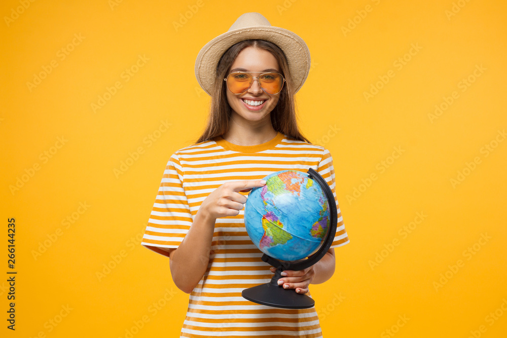 Fototapeta premium Travel agency advertising. Choosing place to be concept. Young female tourist holding globe with one hand and pointing with finger to destination, isolated on yellow background