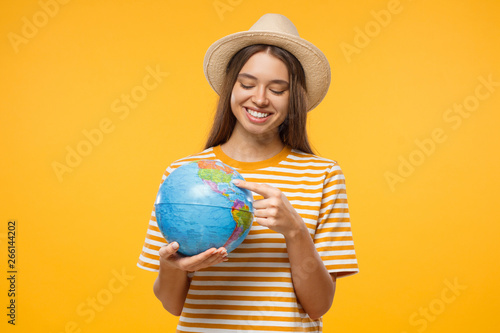  Choosing place to travel concept. Young woman tourist holding globe with one hand and pointing with finger to destination, isolated on yellow background
