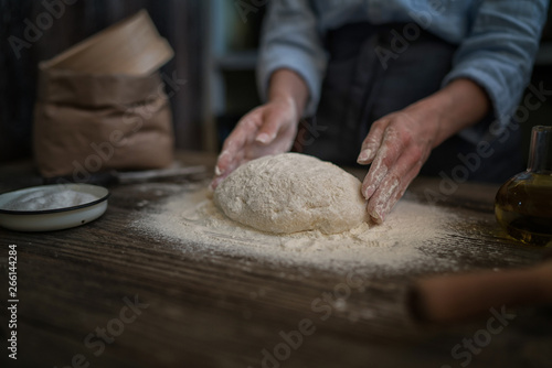 Female hands holding dough in ball shape. Basic homemade dough with ingredients on the side on wooden table with natural light. Home healthy food. Selective focus. Focus on the dough.