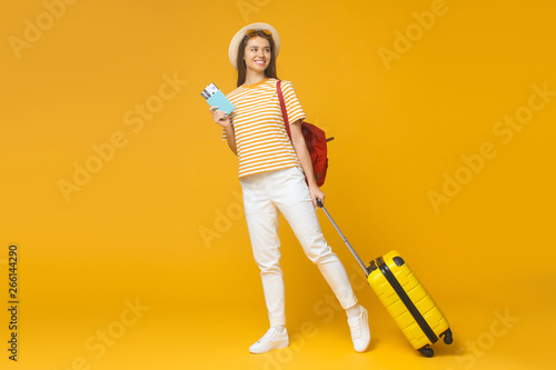 Smiling tourist girl standing with suitcase and backpack, isolated on yellow background. Travel concept
