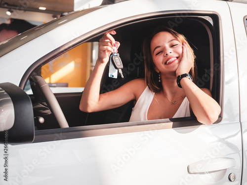 Side view of a beautiful girl smiling and looking at camera. Gorgeous young woman holding keys of her new white auto. Happy female client of car dealership buying automobile © kravik93