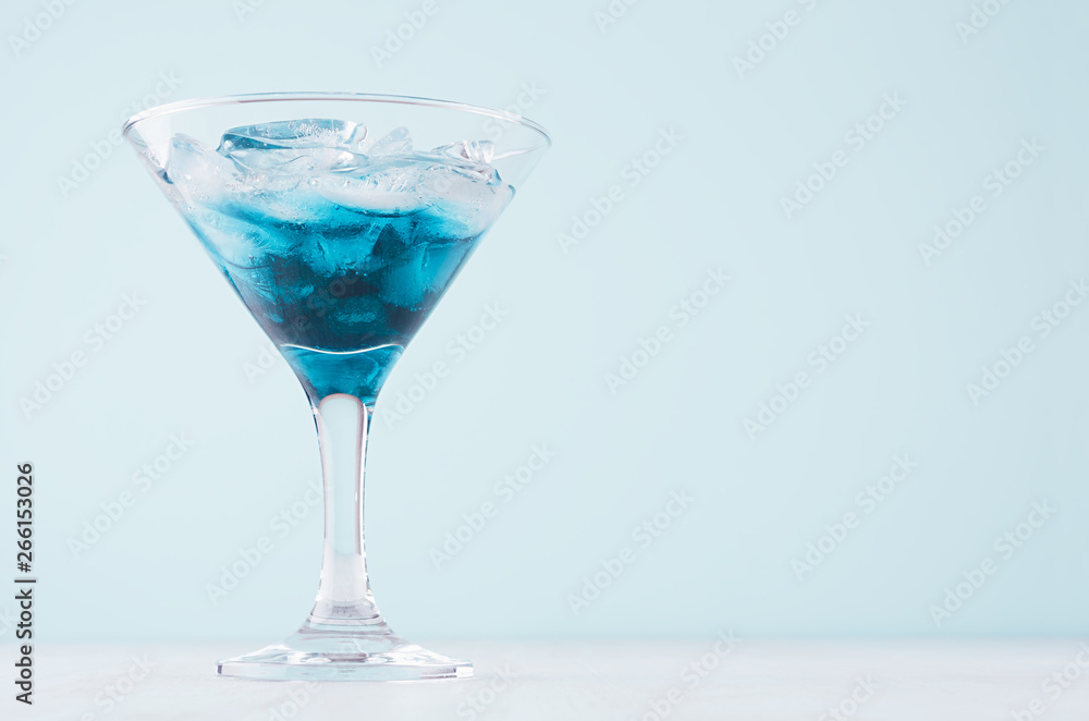 Blue tropical alcohol cocktail with liquor curacao with ice cubes in wineglass on white wood table and blue color wall, copy space.