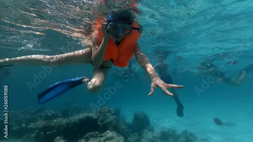 Large group of tourists with scuba masks swim and dive among black and white striped fish near the coral reef. Adult girl in a life jacket swims and looks into the camera. Underwater footage.