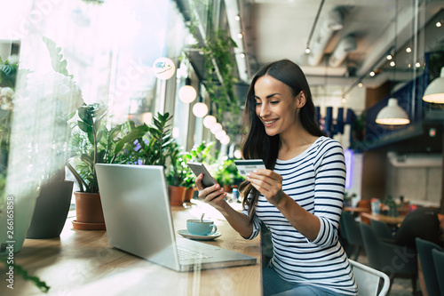 Beautiful smiling young woman using laptop and doing shopping online with help of her smartphone and card