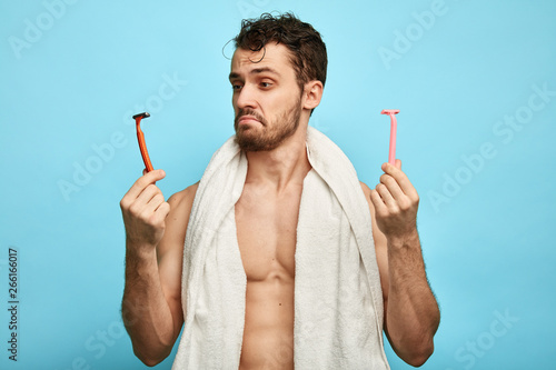 emotional confused muscular man with a towel on his neck holding two shavers. close up portrait