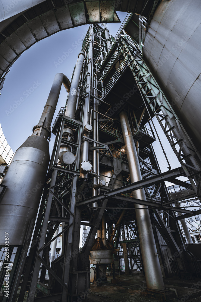 A wide-angle vertical view from the bottom of a modern oil refinery or a contemporary fuel factory facility in an industrial zone, with a round bridge, plenty of pipes, metal beams, tanks, and stairs