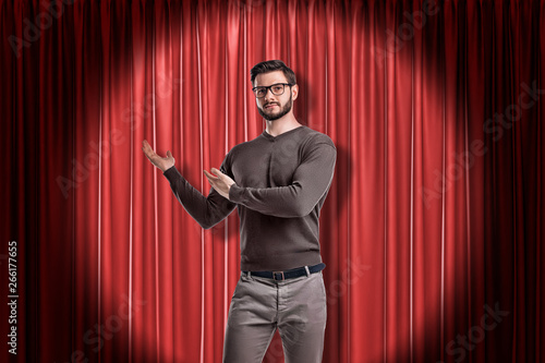 Front crop view of young handsome man in casual clothes and glasses standing in spotlight against red stage curtain and making presenting gesture at it.