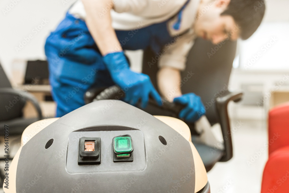 Young man in workwear and rubber gloves cleans the office chair with professional equipment. Young man in workwear and rubber gloves cleans the office chair with professional equipment.