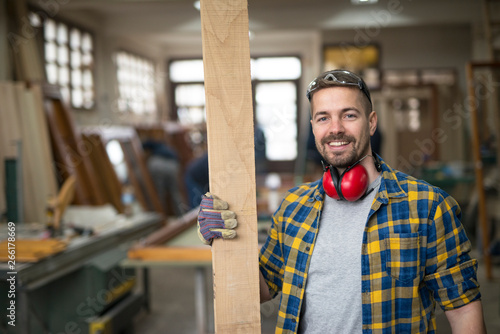 Tableau sur toile Portrait of smiling worker in carpentry workshop holding plank wood material
