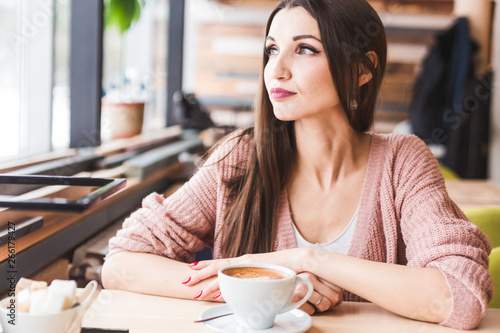 Beautiful young woman sits at a table in a cafe with a cup of coffee and looks out the window