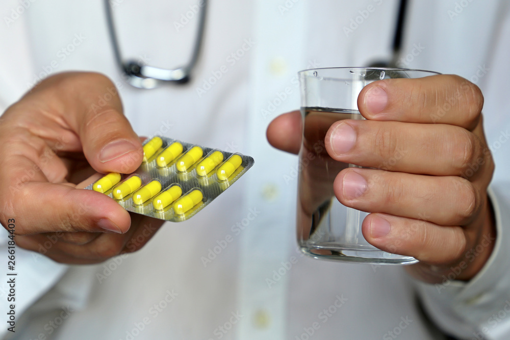 Doctor holding pills and glass of water, therapist giving medication in blister pack with yellow capsules. Concept of medical prescription, dose of drugs, vitamins, pharmacy Doctor holding pills and glass of water, therapist giving medication in blister pack with yellow capsules. Concept of medical prescription, dose of drugs, vitamins, pharmacy