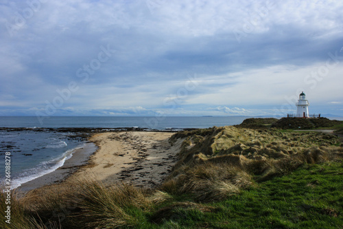 View on a bay with a sandy beach and cloudy sky, white lighthouse in the back, South Island, New Zealand