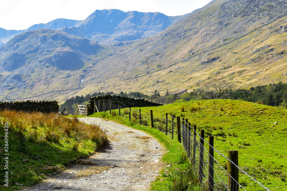 Gravel path leading up to stone wall with wooden gate. Stunning nature in the background. Grisedale pike, Lake District, England, UK