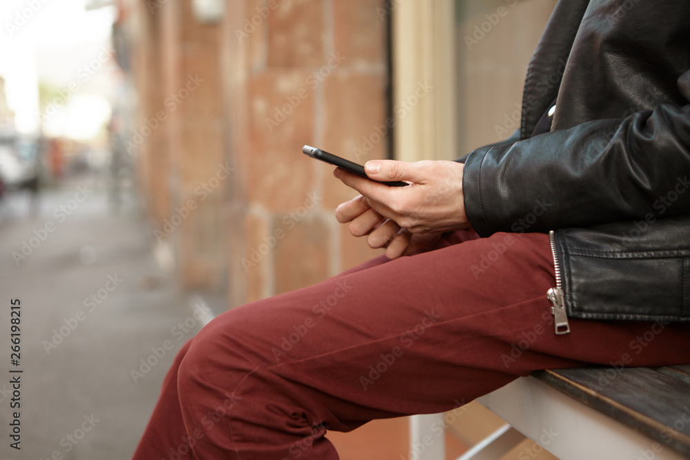 Cropped shot of unrecognizable man in leather jacket sitting outside on bench on empty street holding generic cell phone, chatting with friends, typing text messages using 4g internet connection Cropped shot of unrecognizable man in leather jacket sitting outside on bench on empty street holding generic cell phone, chatting with friends, typing text messages using 4g internet connection
