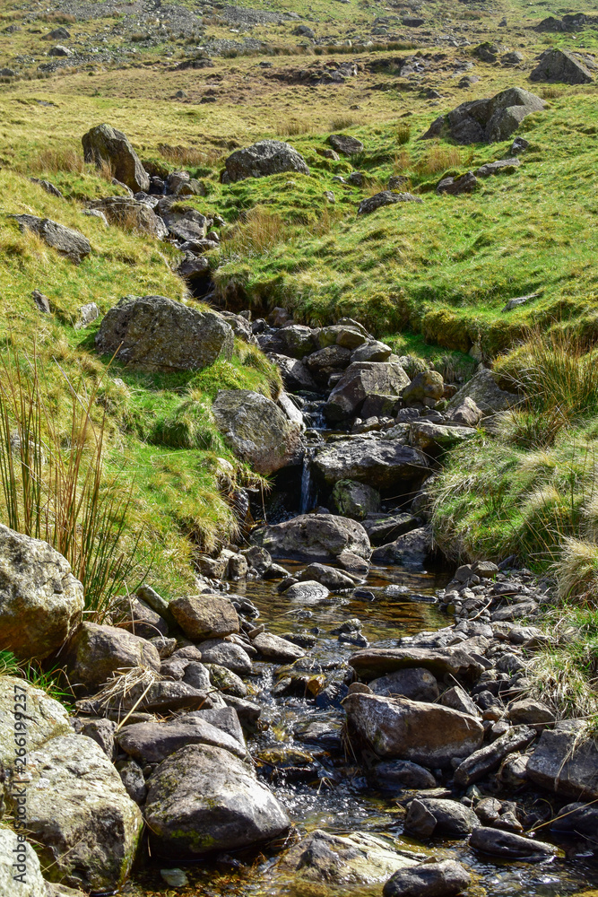 Small stream on dark rocks next to grassy fields. Bright sunny day. Grisedale pike, Lake District, England, UK
