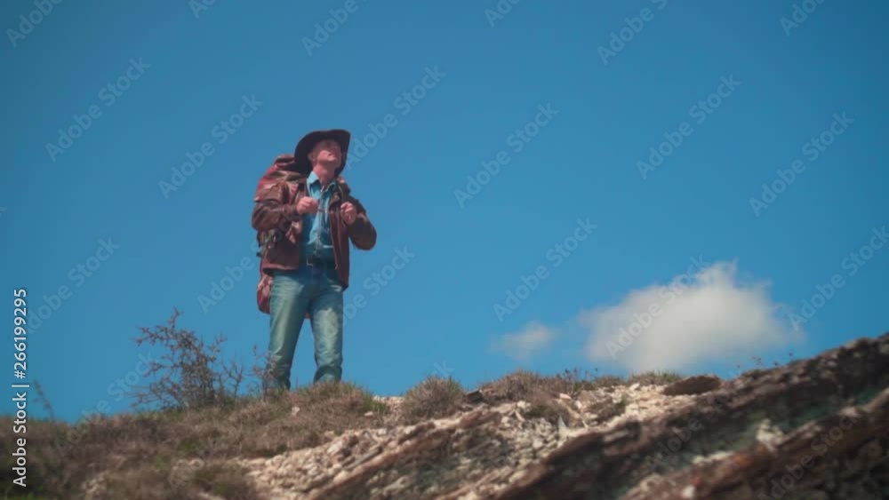 In the mountains, a man in a cowboy hat, leather jacket, blue jeans, a large tourist backpack on his shoulders. The man looks around, taking off his glasses and looking at him.
