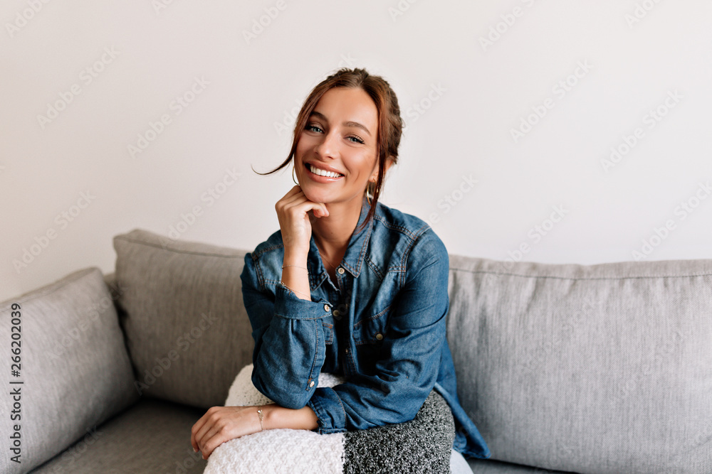 Happy laughing girl with tanned skin and white teeth sitting at sofa at cosy apartment and posing at camera