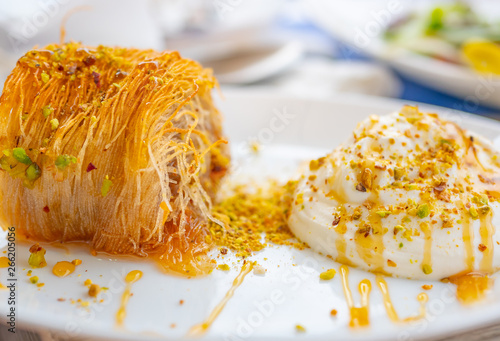 Close up of a succulent Greek dessert. Greek kataifi with honey yogurt and grated pistachios on a white plate and a blurred background. Horizontal view