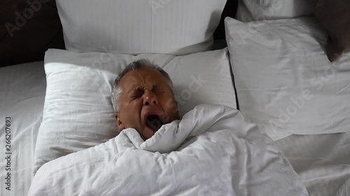 An elderly man wakes up and yawns while lying in bed in the early morning.