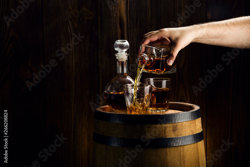 male hand pouring rum in glasses, strong alcoholic drinks in decanters are on the barrel
