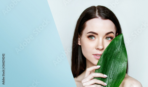 Portrait of young beautiful woman with healthy glow perfect smooth skin holds green tropical leaf, look into the hole of colored blue paper. Model with natural nude make up.