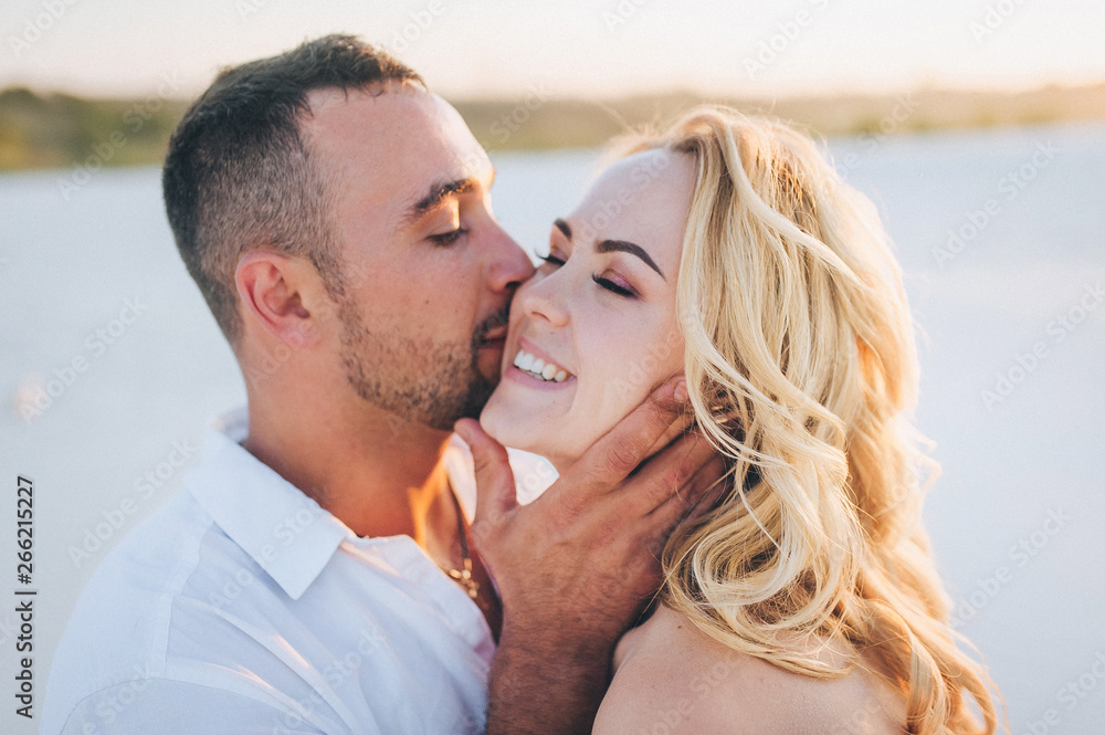 Tanned young guy kisses a beautiful blonde girl against the setting sun close up. Desert, sandy beach, vacation and rest. Sunset. Honeymoon concept.