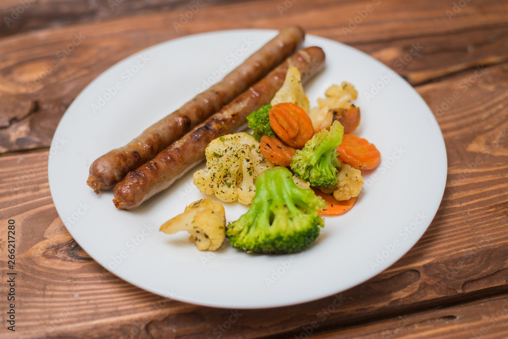 Meat sausages and vegetables fried on a white plate on a wooden background. Meat sausages and vegetables fried on a white plate on a wooden background.