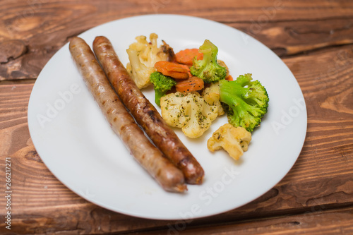 Meat sausages and vegetables fried on a white plate on a wooden background.