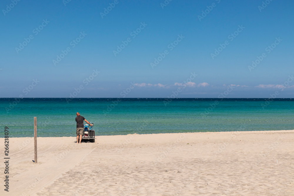 The bay of Torre dell'Orso, with its high cliffs, in Salento, Puglia, Italy. Turquoise sea and blue sky, sunny day in summer. In a bathhouse, a man cleans the sand with a small tractor (sand cleaner).