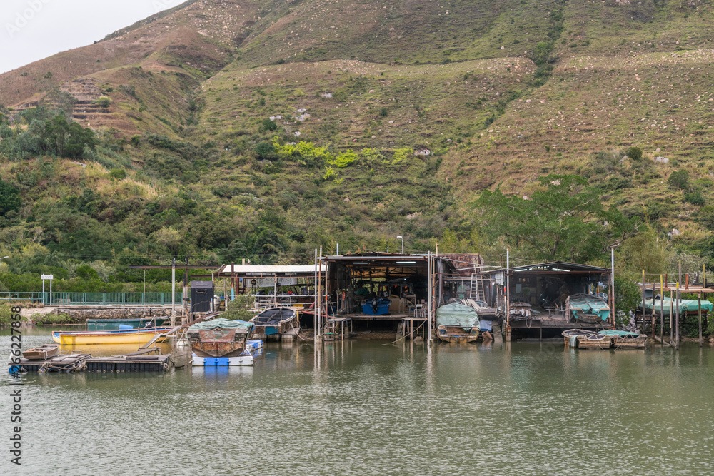 Hong Kong, China - March 7, 2019: Fisherman house on stilts on Tai O River with dry docks to park the sloops and green large hill in back. Color added by tarps on boats.