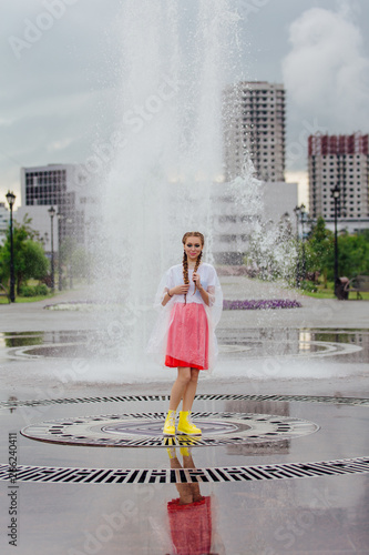 Young wet pretty girl with two braids in yellow boots and with transparent umbrella stands inside of fountain.