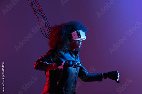 Mod curly dark haired girl dressed in black leather jacket and gloves uses the virtual reality glasses on her head in the dark studio with neon light