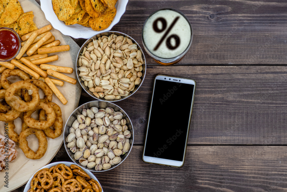 Mix of snacks and beer with percent sign on a beer foam, and smartpnone on dark wooden background. Internet delivery concept. Top view. Empty space for text Mix of snacks and beer with percent sign on a beer foam, and smartpnone on dark wooden background. Internet delivery concept. Top view. Empty space for text