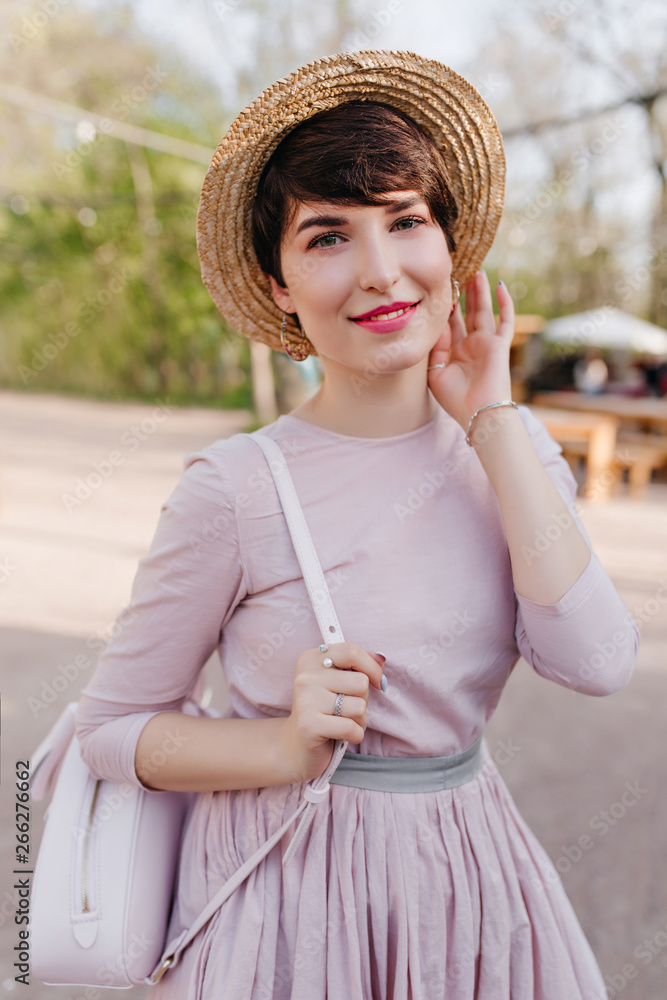 Lovely young woman with shiny short hair posing with pleasure during walk in park. Portrait of trendy girl wearing vintage attire and silver jewelry, standing outside with white backpack.