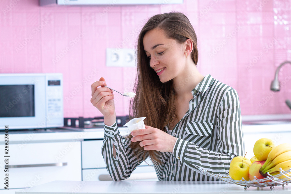 Portrait of a young, smiling, happy woman holding yogurt cup and eating fresh yogurt for snack at home in the kitchen Portrait of a young, smiling, happy woman holding yogurt cup and eating fresh yogurt for snack at home in the kitchen