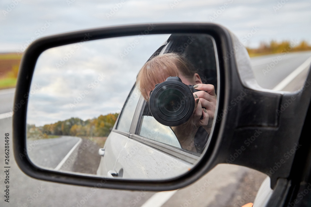 Car mirror, the reflection of the female photographer in it and landscape with field and tree