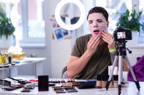 Self-confident dark-haired guy in greet t-shirt applying moisturizing face mask