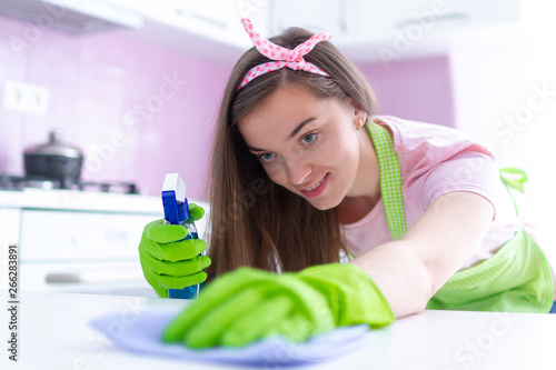 Young attractive housewife in protective rubber gloves and apron wiping dust off furniture at kitchen at home using a rag and spray. Spring cleaning and housekeeping