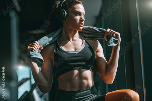 Young attractive strong muscular female bodybuilder with ponytail and headphones posing in gym with towel around neck. Work out in silence, let your success be your noise.