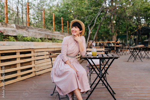 Excited brunette girl in beautiful old-fashioned dress touching face with charming smile resting in outdoor cafe. Portrait of joyful young woman in vintage attire drinks juice in open-air restaurant.