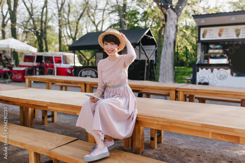 Smiling girl in long dress and trendy shoes sitting on wooden table holding mobile phone in hand. Portrait of lovely young woman wearing straw hat, resting in open-air restaurant with snack-bars.