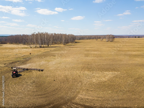 Aerial of Red tractor with a trailed plow for mowing and weeding fields for the agro-industry of yellow color under the blue sky, a clear spring day. Preparation for planting crops rural technology.