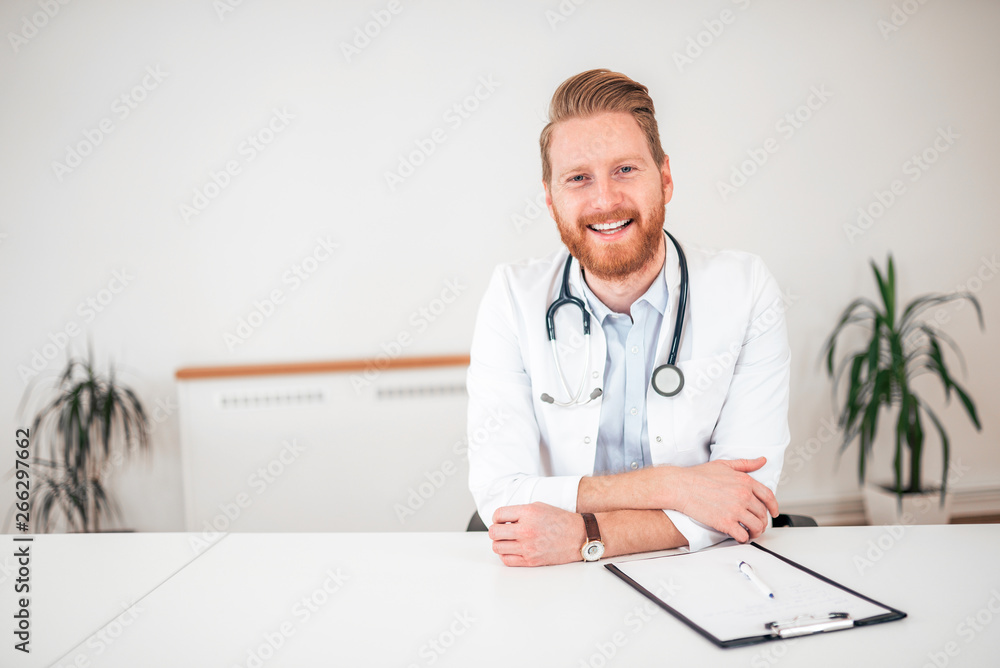 Portrait of a smiling ginger general practitioner sitting at office desk. Portrait of a smiling ginger general practitioner sitting at office desk.