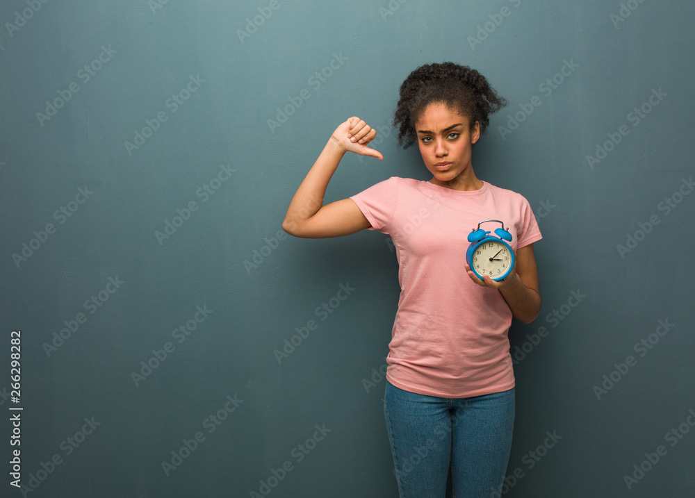 Young black woman pointing fingers, example to follow. She is holding an alarm clock.